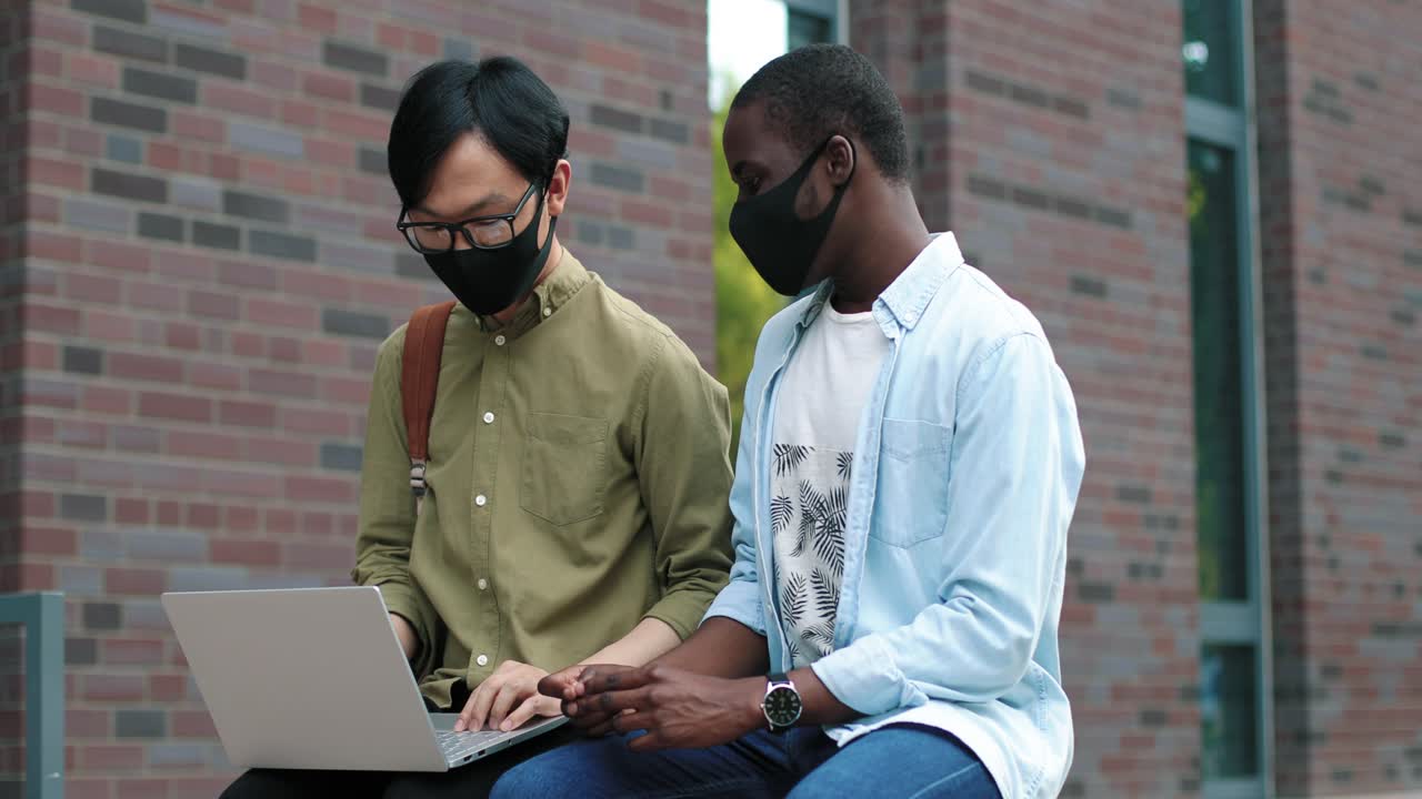 Waist up portrait view of asian and african american male students wearing protective masks sitting in the street and looking at the laptop while studying new the college