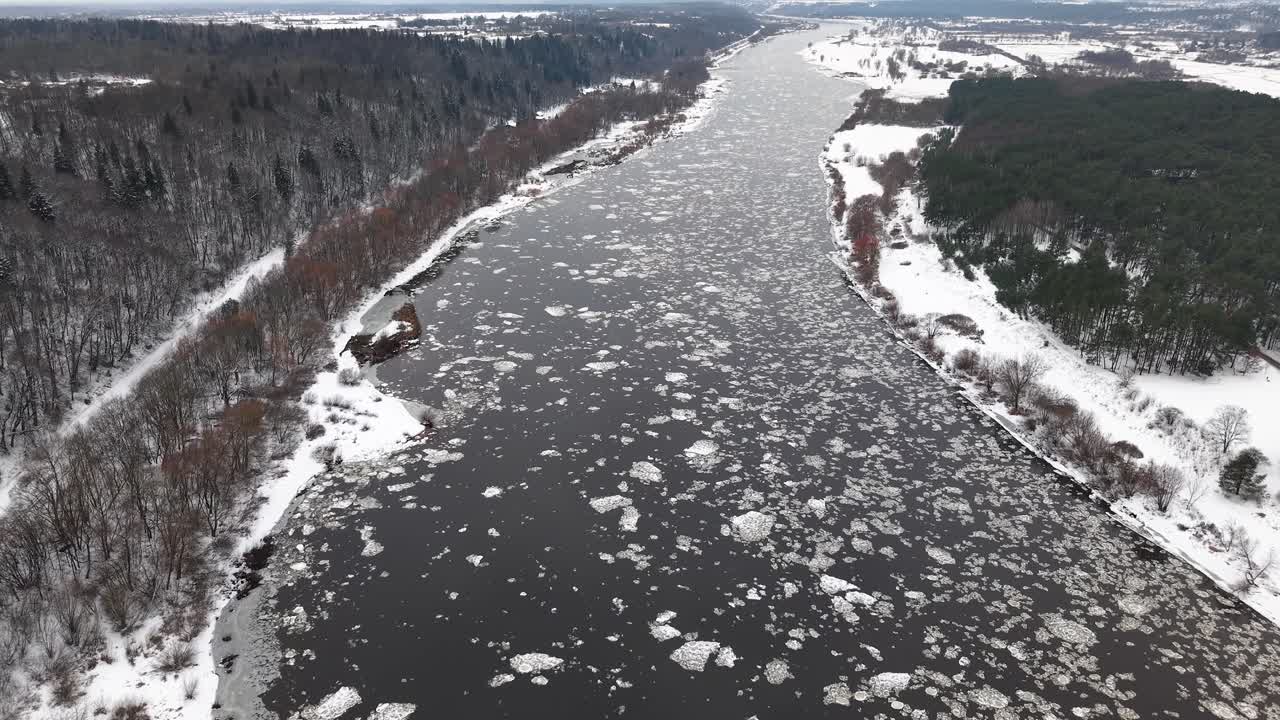 Ilmakuva, jossa jäätynyt joki kiemurtelee lumisen maiseman läpi. Jäälohkareet ajelehtivat vedessä, jota reunustavat kuuraiset puut ja talvinen maasto seesteisessä, kylmässä maisemassa.