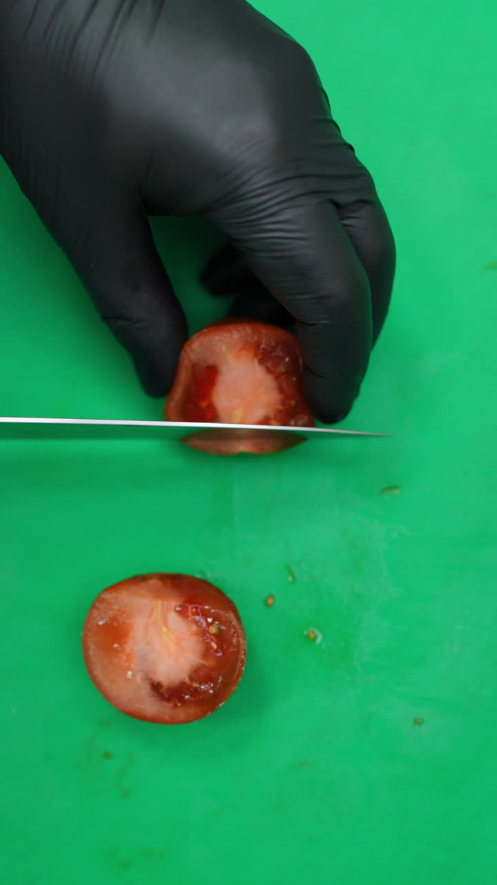 Vertical footage Gloved hands skillfully slice a ripe, red tomato into several pieces on a green cutting board, demonstrating precision and culinary expertise