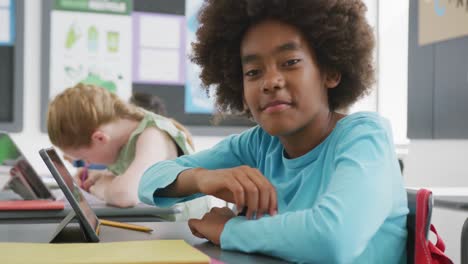 Video-portrait-of-smiling-african-american-schoolboy-at-desk-using-tablet-in-class,-copy-space