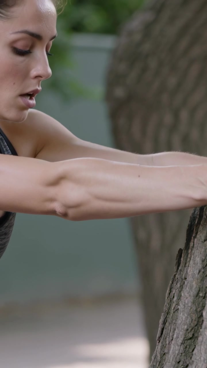 Video verticale: La telecamera mette a fuoco una donna in forma che preme le braccia contro un albero in giardino per fitness in canotta