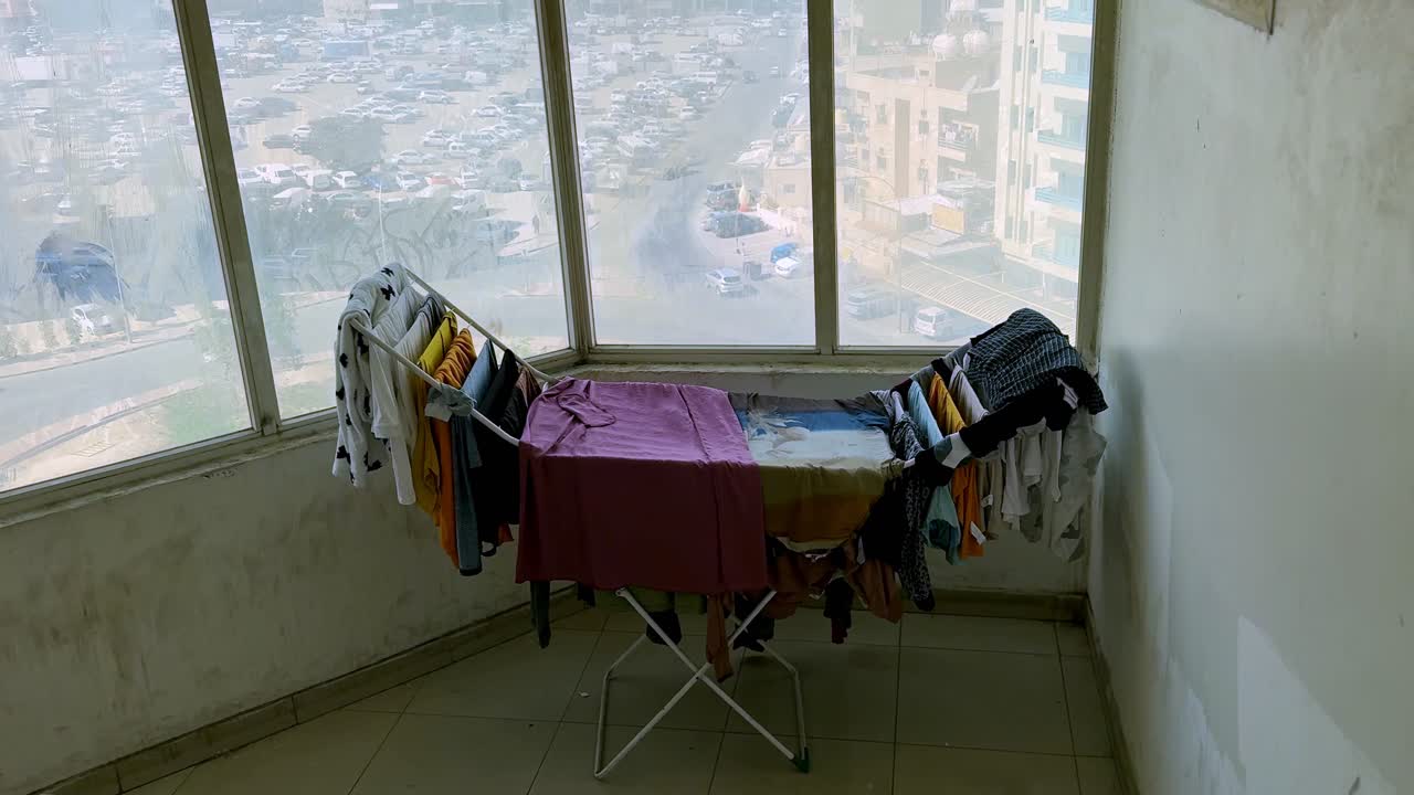 Wide interior shot of a foldable drying rack covered with laundry beside large apartment windows overlooking a busy city street. Daylight urban scene showing everyday home routine