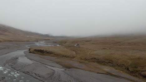 Foggy-river-scenery-and-abandoned-house-not-far-from-shore,-Iceland