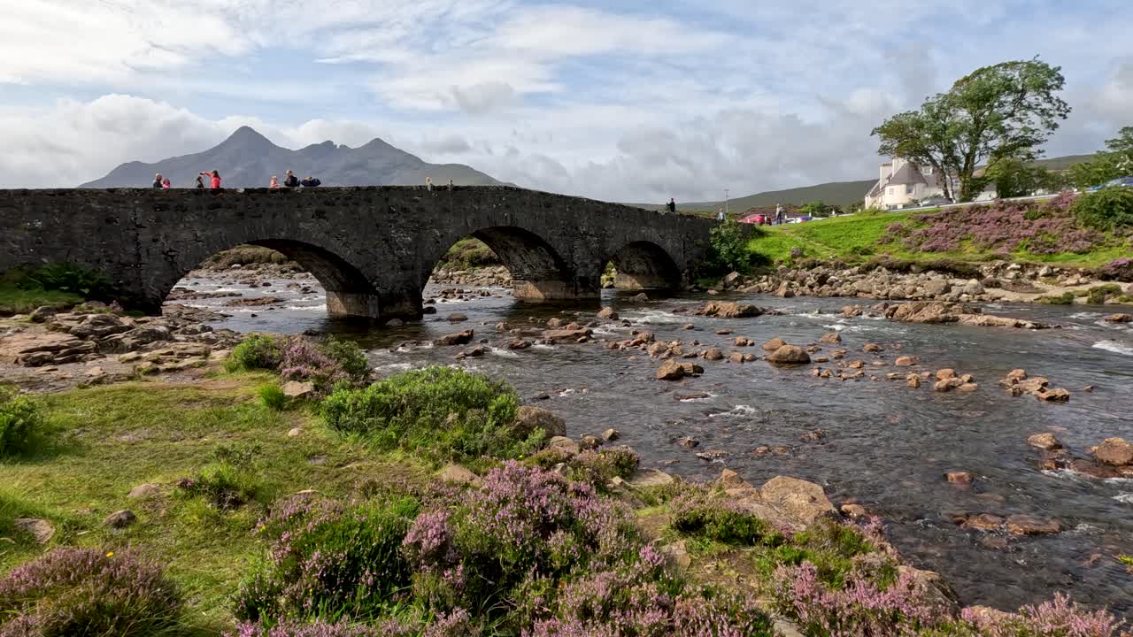 Wide shot of a historic stone bridge crossing a rocky river, with distant mountains and lush greenery under soft daylight. Subtle camera pan reveals scenic landscape