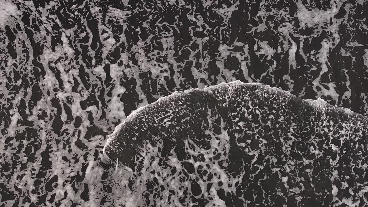 Top-down aerial view of foamy waves crashing onto the black sand.