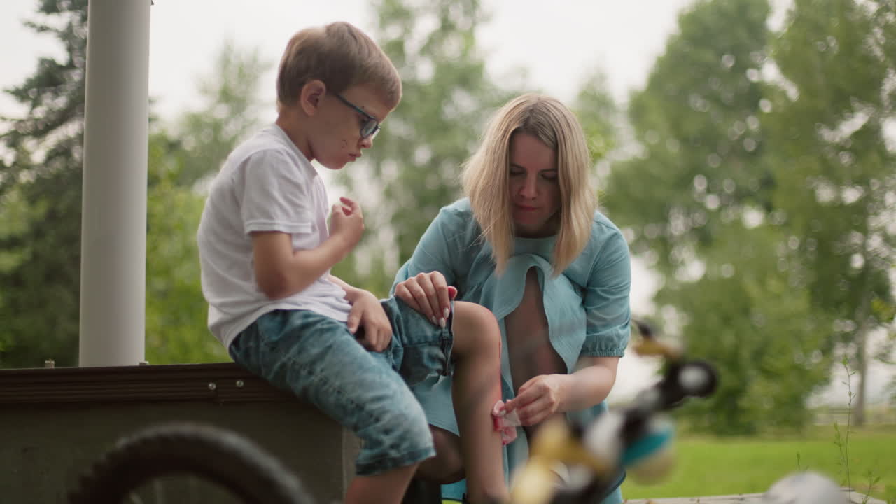 una madre se agacha mientras limpia cuidadosamente la pierna sangrante de su hijo, el niño parece molesto mientras está sentado en un banco