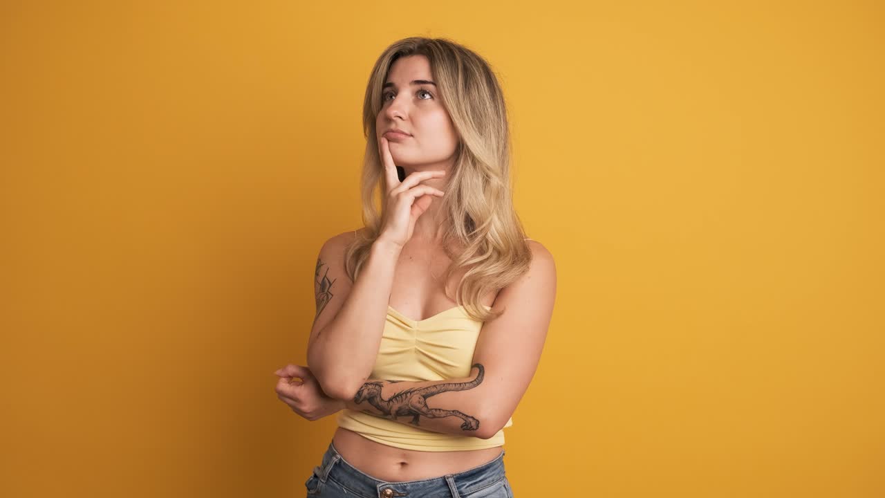 Thoughtful young woman standing with hand at chin on yellow background