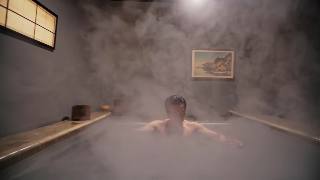 A man relaxes in a steaming hot tub, arms outstretched. The video captures a serene, cinematic
