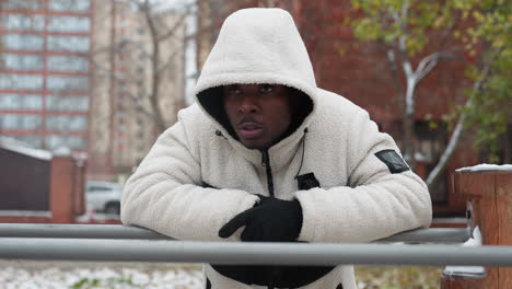 music artist in white hoodie and gloves rests on iron bar outdoors, cold air visible from mouth, background features urban buildings, snowy ground, and trees