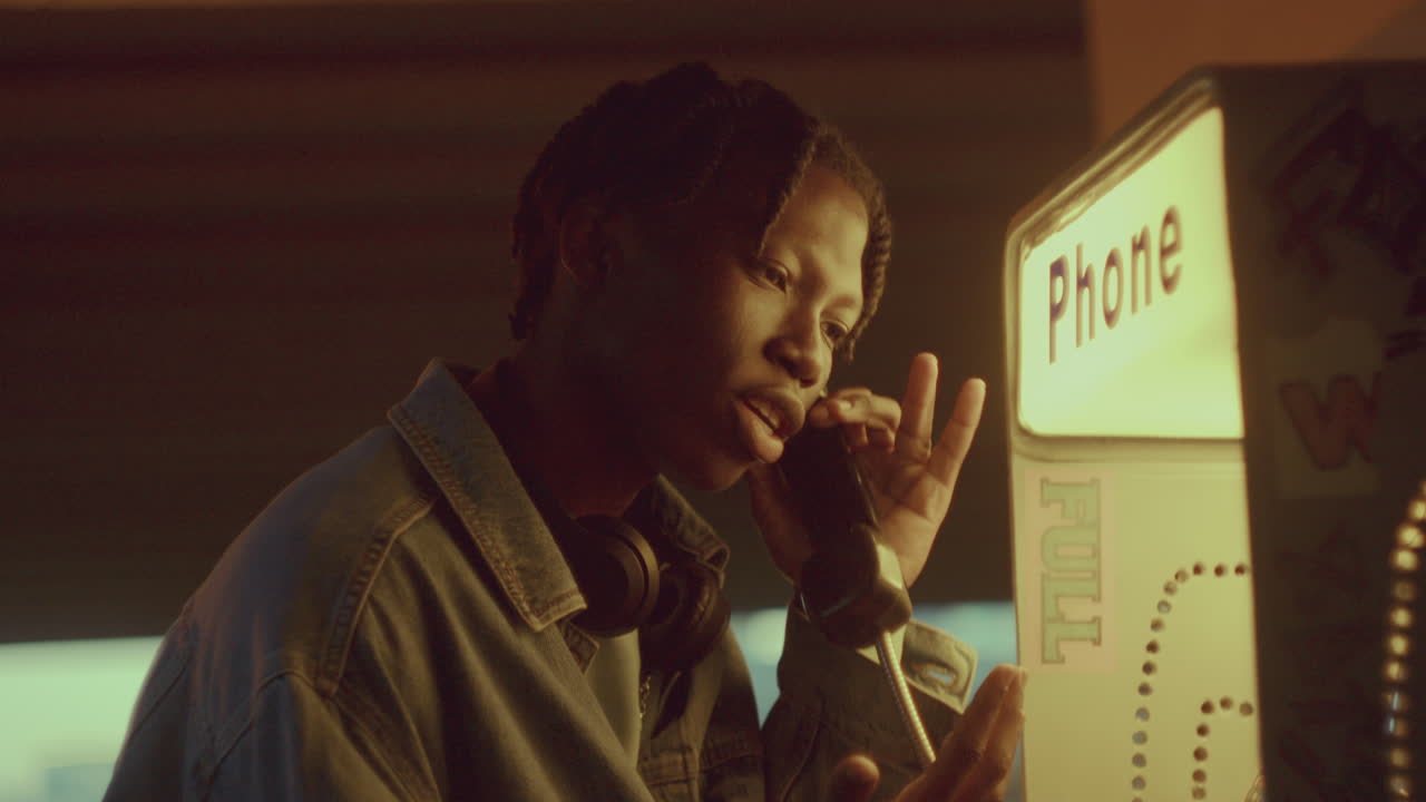 Young Black Man Talking Expressively on Street Payphone in the Evening