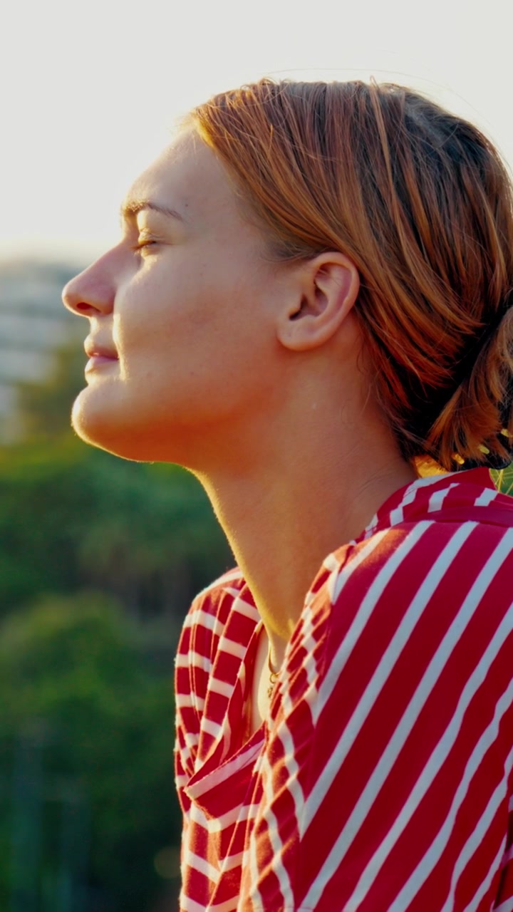 Video verticale in primo piano di una giovane donna con una camicia a righe rosse, a occhi chiusi, che si gode pacificamente la luce del tramonto all'aperto. Ideale per i social media.