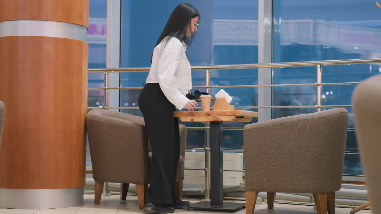 Young girl walks toward cafeteria seating area and gently drops coffee cup and gadget on wooden table near glass wall reflecting outdoor environment before preparing to sit on cozy armchair