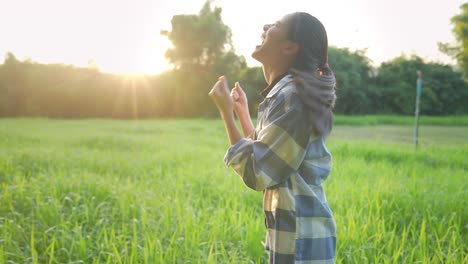 a happy successful female farmer  celebrating herself with cheerfully dance with raising fists up while standing against on a growth green crops field and beautiful golden sunset, excited celebrating