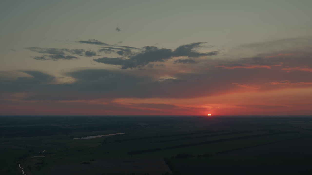 Rising sunset sky with dramatic dark dusk clouds forming surreal shapes under vibrant red and orange glow while sun casts subtle lens flare over expansive flat farmland horizon with distant trees