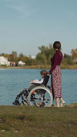 Mother and daughter in wheelchair walk on shore of lake. Young woman touches child hair gently. Loving mom and kid with disability enjoy river view