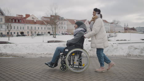 side view of happy muslim woman taking her disabled friend in wheelchair on a walk around the city in winter