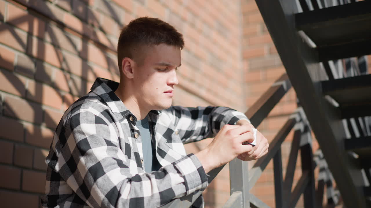 white man resting on iron railing retrieves cigarette from pack holds it to lips placing it in mouth while leaning on metal railing wearing checkered shirt under sunlit brick staircase backdrop