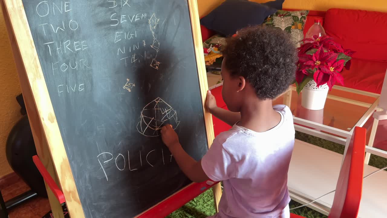 A 5-year-old cute black kid sitting on a red chair drawing on a chalkboar at home. He is wearing curly hair and a white shirt.