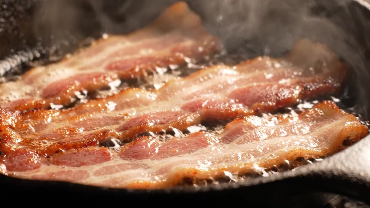 Strisce sfrigolanti di pancetta che friggono in una padella di ghisa, mettendo in mostra la deliziosa consistenza croccante e il colore dorato esaltati dal processo di cottura.