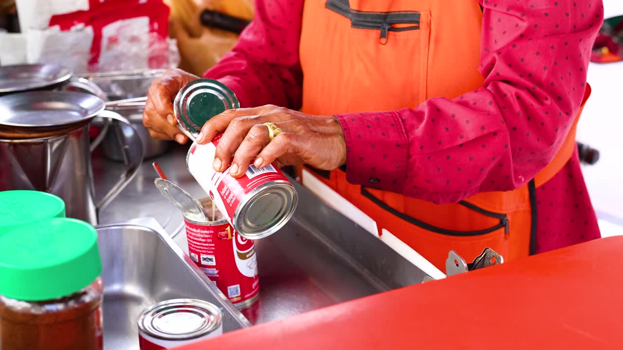 A person in an orange apron pours condensed milk into a container, preparing tea in a vibrant kitchen setting