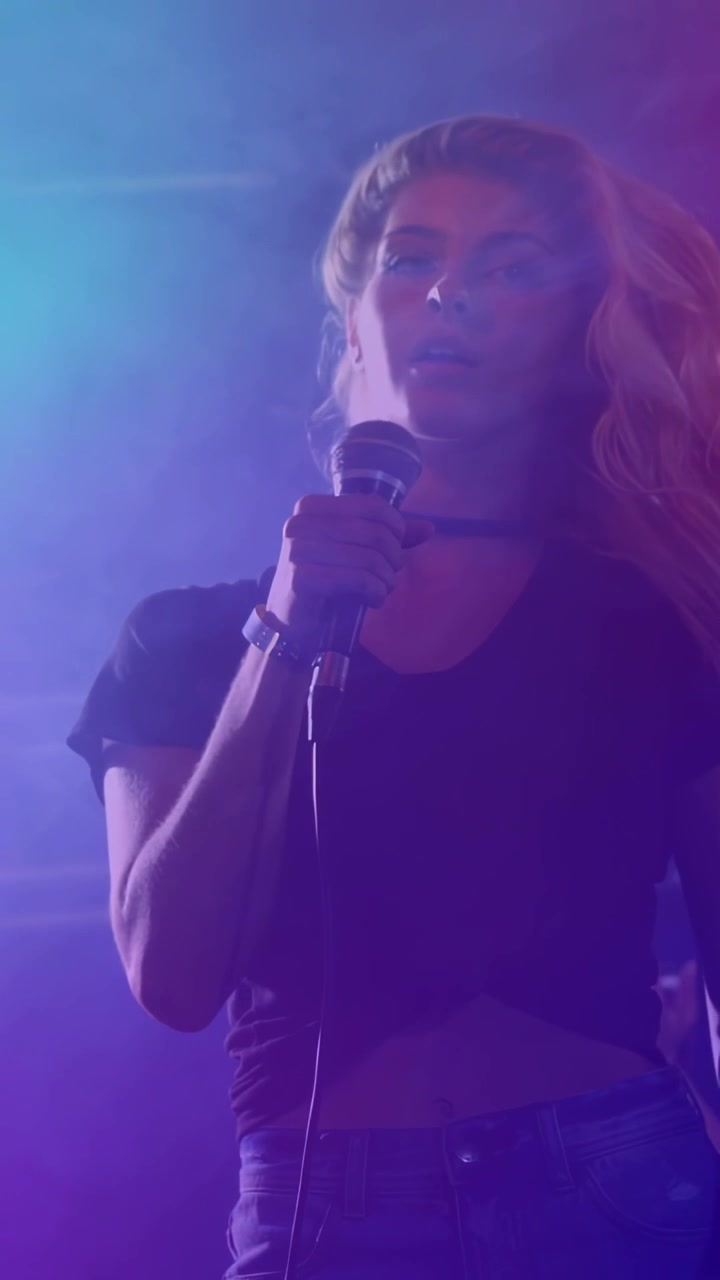 Vertical video: Female singer holding wired mic, starting long note, performing music in blue haze