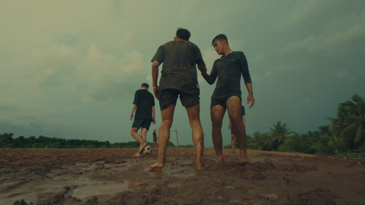 A football player fouls another in a muddy field, with a teammate helping up in slow motion