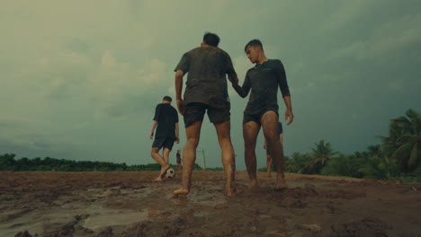 a football player fouls another in a muddy field, with a teammate helping up in slow motion
