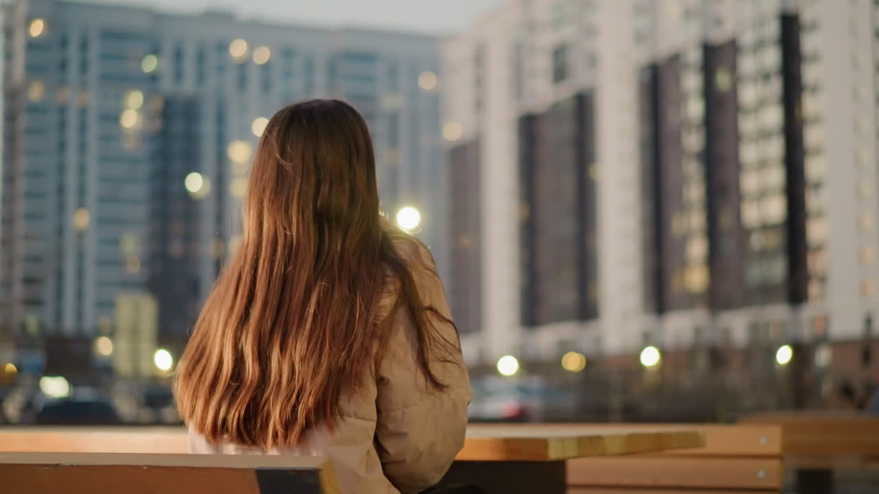 도시 공원의 벤치에 앉아있는 긴 갈색 머리카락을 가진 복아 재을 입은 여자의 뒷.