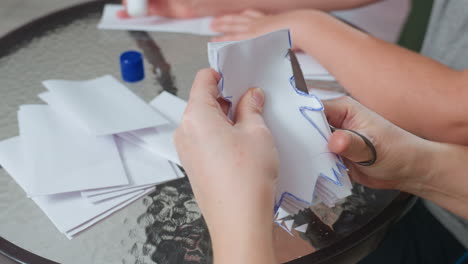 hands engaged in creative process cutting white paper with scissor forming craft design while another hand applies glue on paper on glass table surrounded by scattered paper pieces