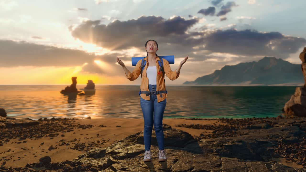 Corpo intero di escursionista asiatica con zaino da alpinismo che allarga le braccia e si gode il panorama mentre è in piedi sulla spiaggia durante il tramonto