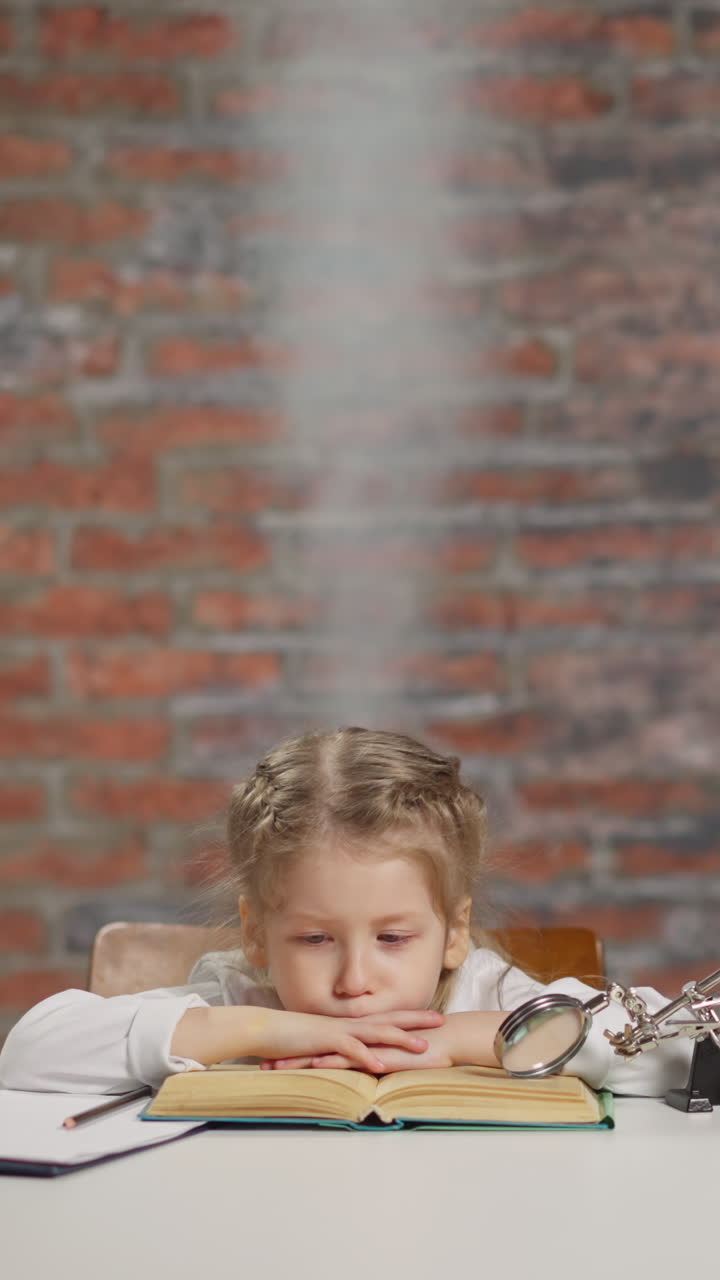 Tired little girl student with steam rising above head with boiled brains reads textbook near magnifying glass and laptop at white table in light studio
