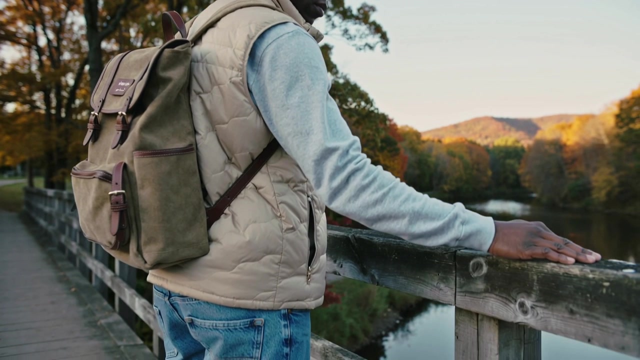 Pausing man beige vest tan backpack on wood bridge sliding hand railing watching river, copy space