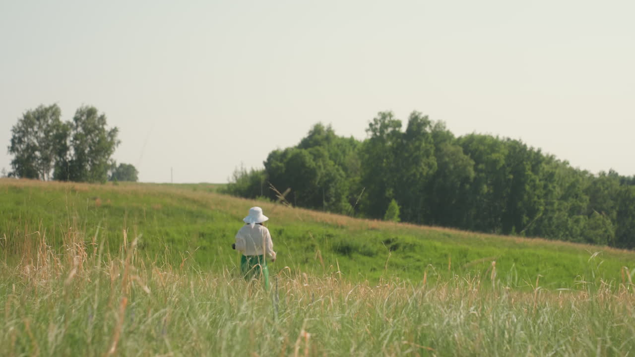 Back view of woman in white hat and green skirt walking alone through tall grassy meadow toward cliff edge, surrounded by soft sunlight and lush green landscape with trees in background