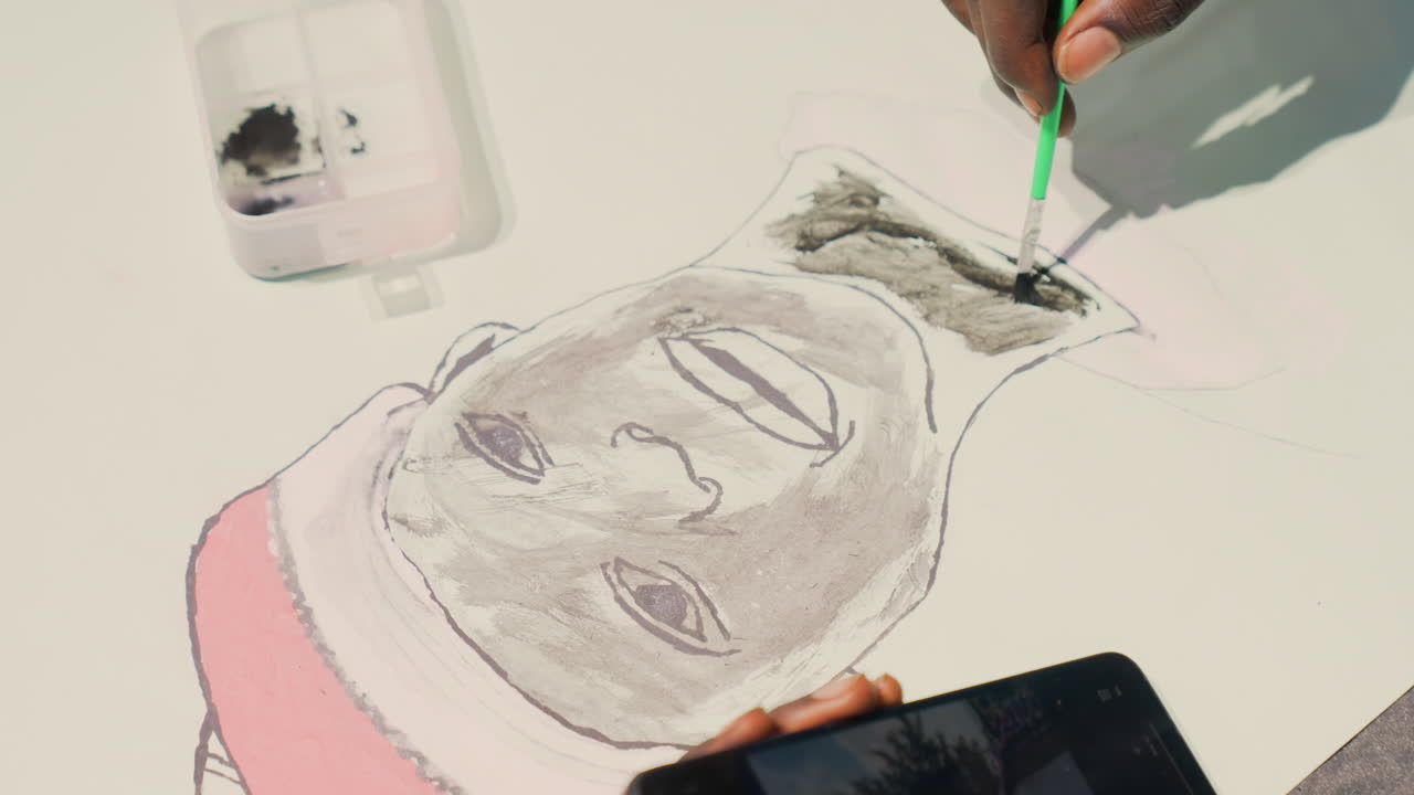 Primer plano de una mano pintando un retrato oscuro de un sujeto afrodescendiente en papel, el artista aplica tinta a la línea del cabello con un pincel pequeño y una herramienta verde, un recipiente de plástico con pigmento, contorno de una gorra rosa, referencia del smartphone.