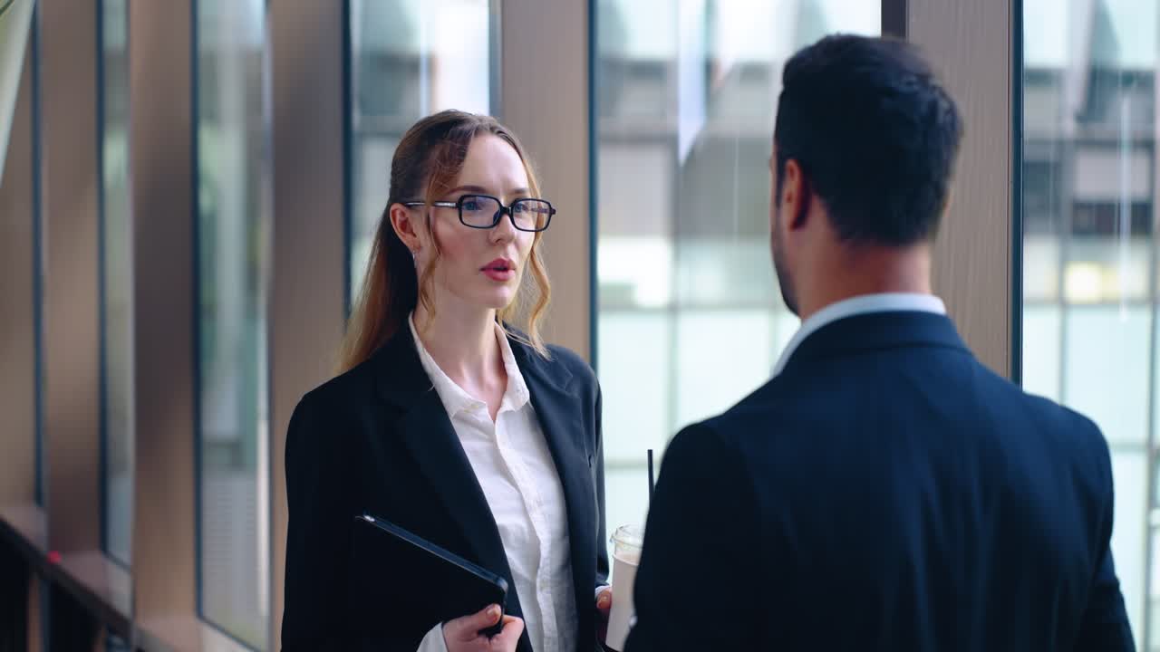 Two office workers greeting each other in hallway with friendly conversation, focus on woman holding drink