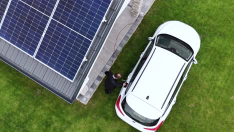 aerial top down of electric white car being charged at home with solar panel installed on rooftop of house