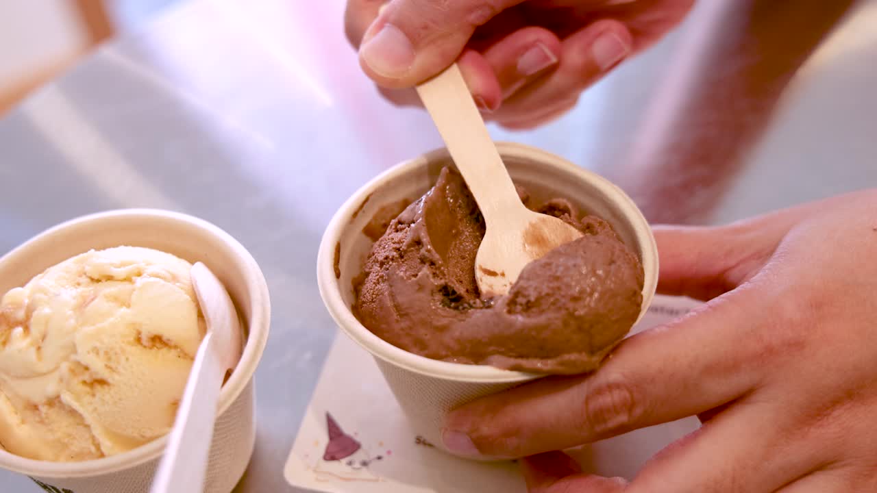 Una mano usa un cucchiaio di legno per prelevare gelato al cioccolato da un bicchiere di carta accanto a gelato alla vaniglia, sotto una luminosa illuminazione interna con una prospettiva ravvicinata