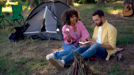 Happy-hiker-couple-ready-to-to-roast-marshmallows-on-a-bonfire.-Caucasian-man-book-and-young-black-female-camping-in-the-forest.