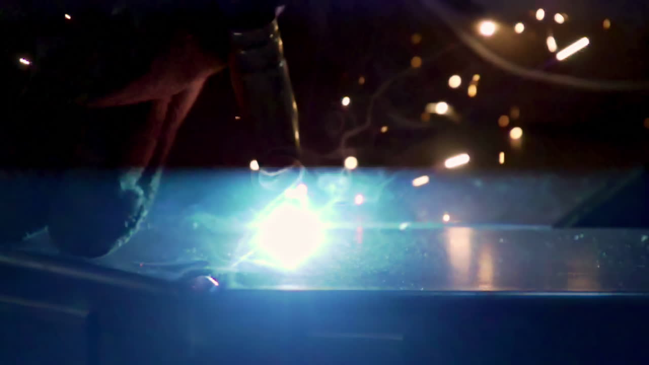 Bright welding sparks and blue smoke rise during metalwork in a dark industrial setting
