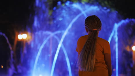 The-child-admires-the-fountain-with-lighting,-sits-on-the-edge-of-the-fountain,-rear-view