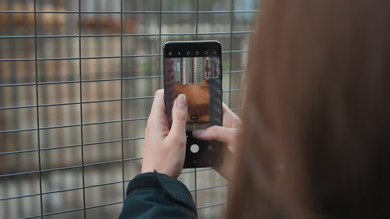hands holding smartphone against metal fence capturing blurred urban scene through grid person composes shot of textured background, tapping screen to focus with shallow depth and overcast light