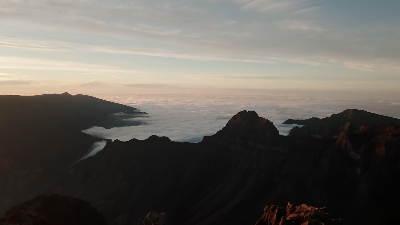 Wide aerial shot of jagged peaks and soft cloud layers spilling through valleys at Pico do Arieiro, Madeira, during early sunrise.