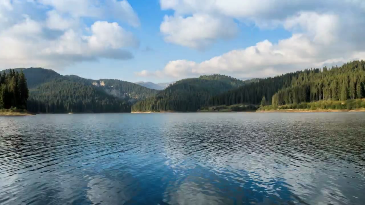 Vue large d'un lac de montagne calme entouré de collines boisées sous un ciel bleu vif et des nuages dérivants. De légères ondulations de l'eau reflètent le paysage paisible.