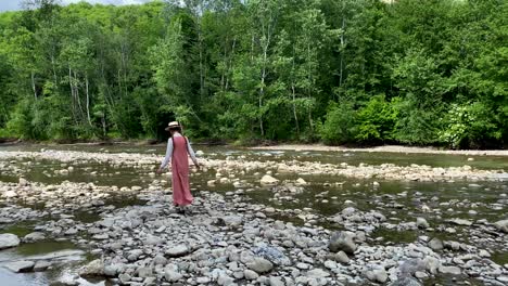 retro styled young woman in straw boater hat and long dress walking on river rocks near forest. romantic girl. natural scenic landscape. summer vacation. vintage aesthetic lifestyle. 4k video