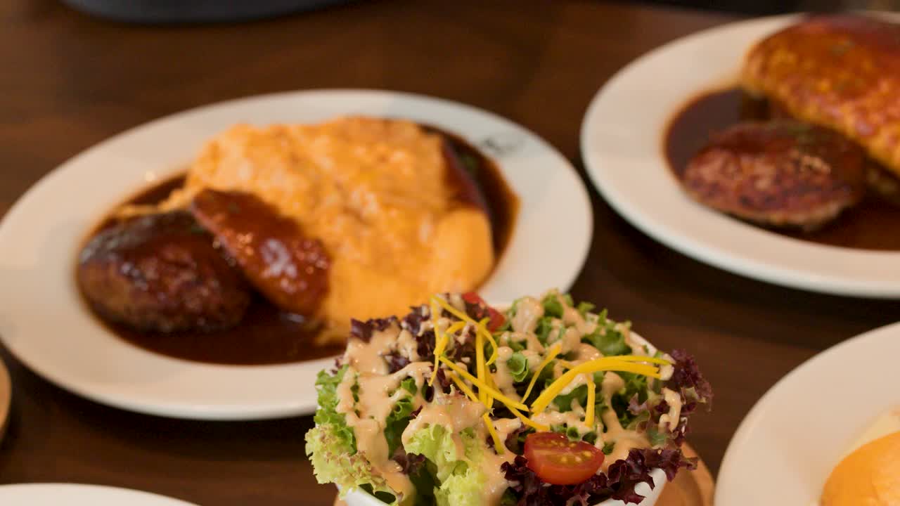 Overhead camera rotates above Japanese omelette rice, salad, soup, and creamy sauce plates