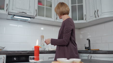 una mujer con un elegante vestido prepara la mesa de almuerzo en una cocina acogedora, una vela roja ardiente en primer plano añade calor al entorno, armarios modernos con iluminación debajo del armario