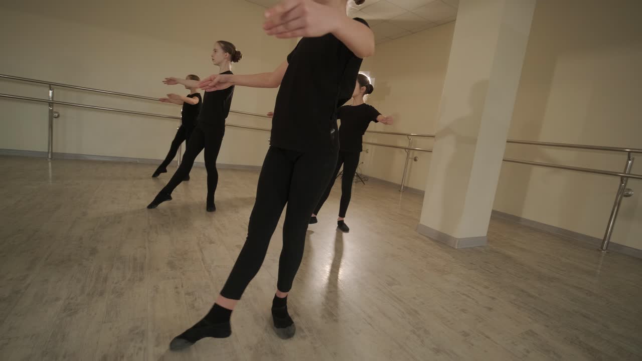 группа молодых студентов балета в черной танцевальной одежде практикует позиции в просторной балетной студии с деревянным полом и настенными решетками. сосредоточенные выражения и синхронизированные движения.