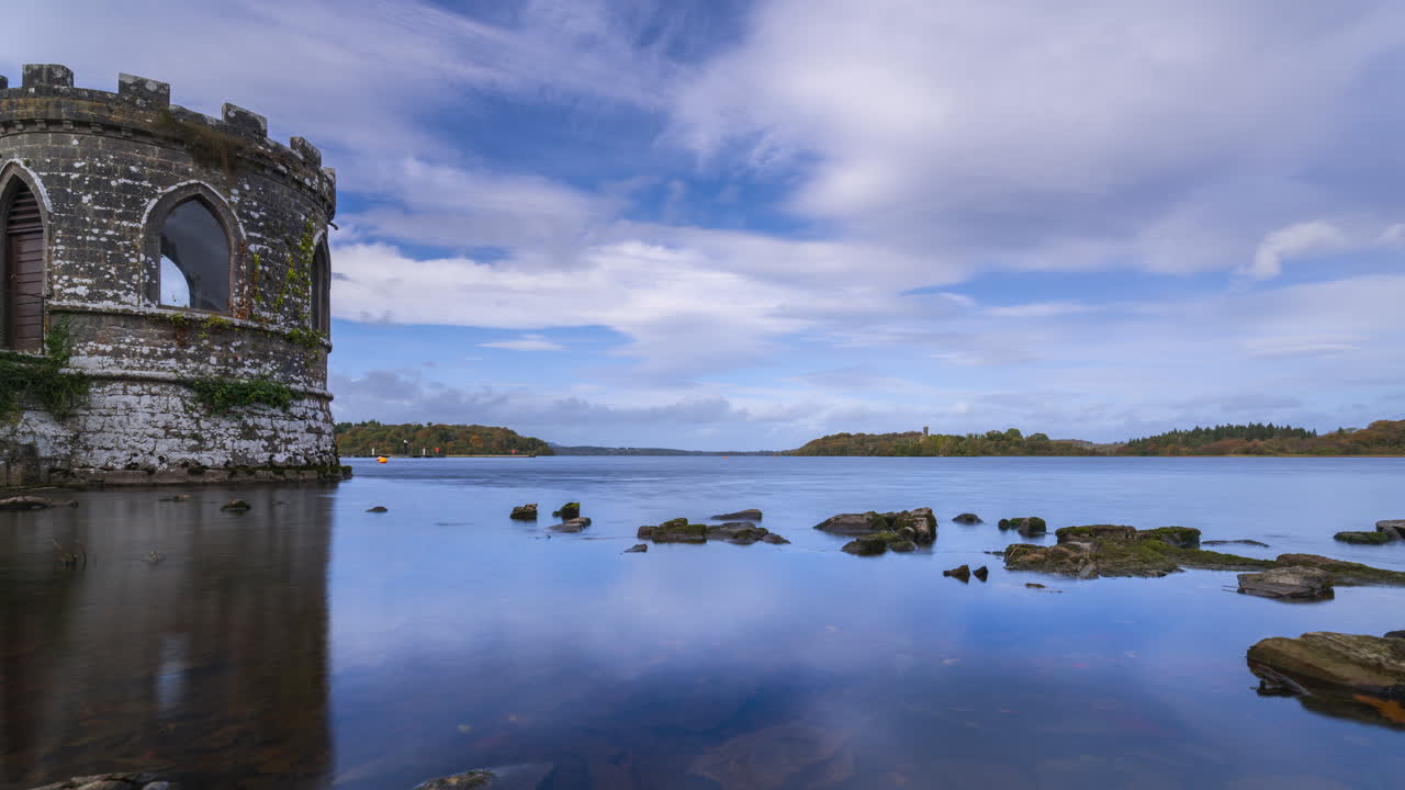 Timelapse av et middelaldertårn ved en innsjø med strandsteiner i forgrunnen og middelalderborg i det fjerne på en delvis skyet dag i Roscommon fylke i Irland