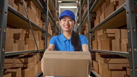 mensajera asiática en uniforme azul mostrando un cartón a la cámara y sonriendo mientras lo entrega en el almacén