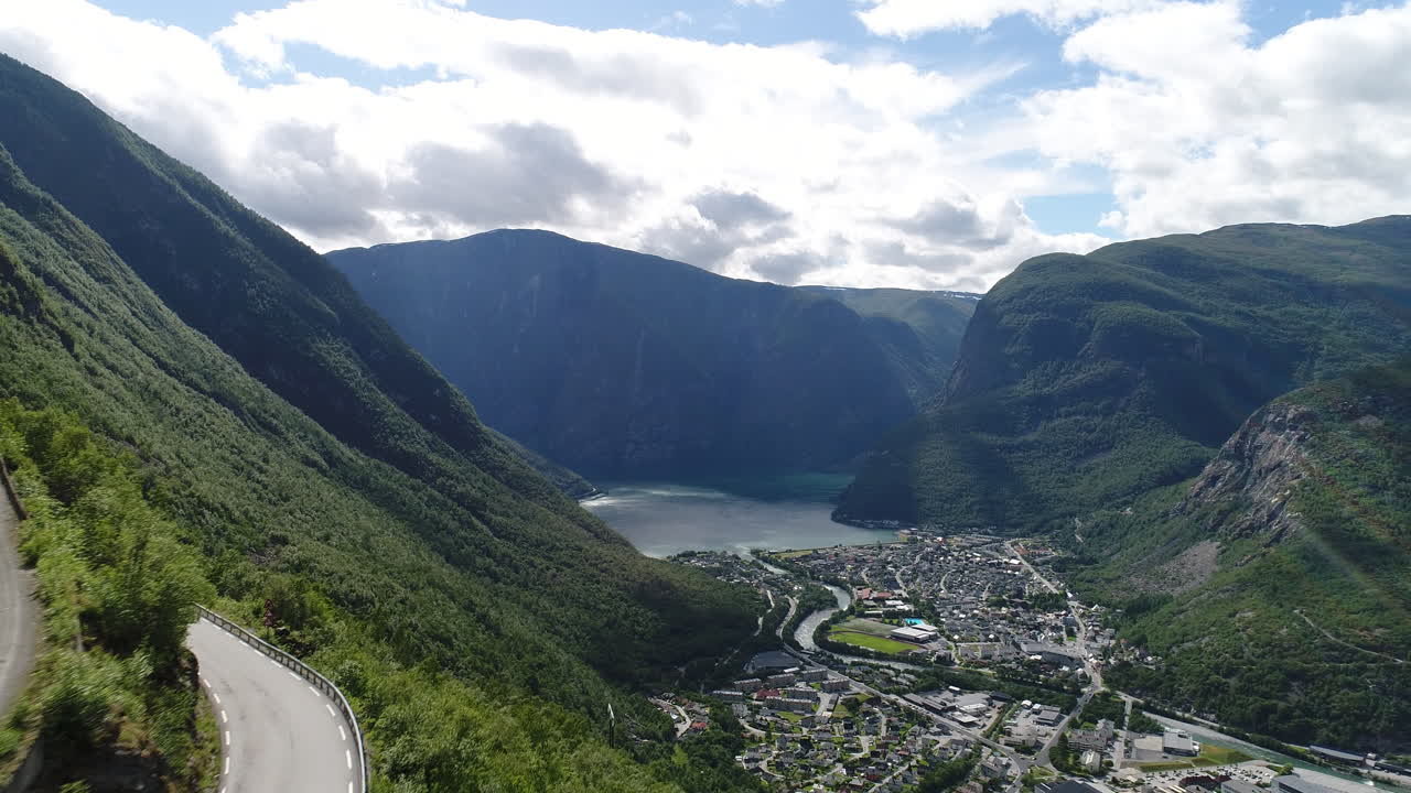 드론은 울불한 푸른 산이 있는 피오르드 의 계곡 마을로 내려가는 휘어지는 길을 보여줍니다.
